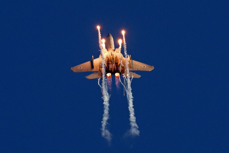 شراره‌ها و رگه‌های دود از یک جنگنده اسرائیلی اف-15 عقاب در حال پرواز در برابر آسمان آبی گسترش یافته است.
